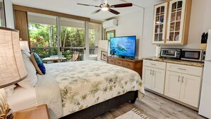 Ein Schlafzimmer mit einem Bett, einem Fernseher, einem Deckenventilator, einem Fenster mit Blick ins Grüne und einer Küchenzeile mit einer Mikrowelle.