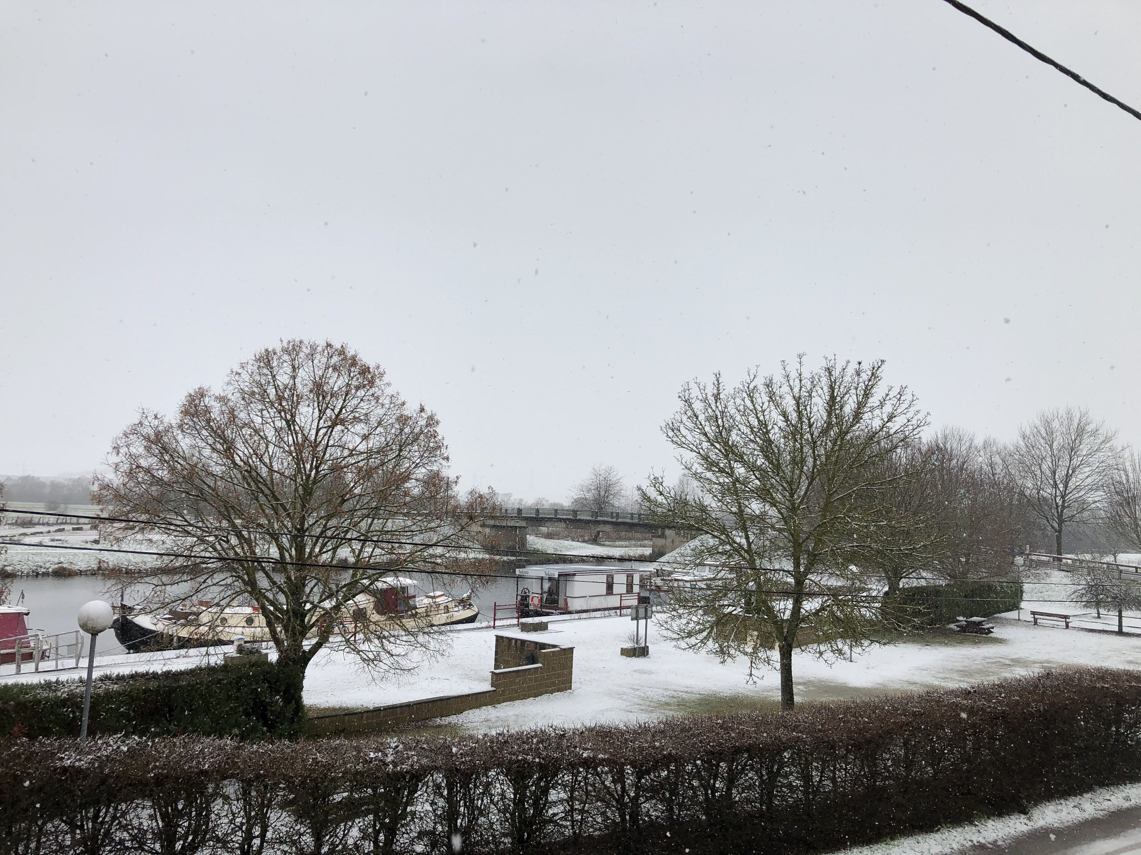A snow-covered landscape with a bridge, trees, and a body of water.
