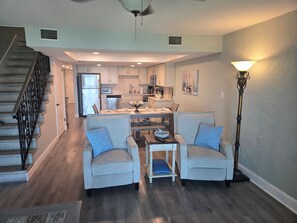 A spacious living area with a dining table, two armchairs, a staircase, and a kitchen in the background.