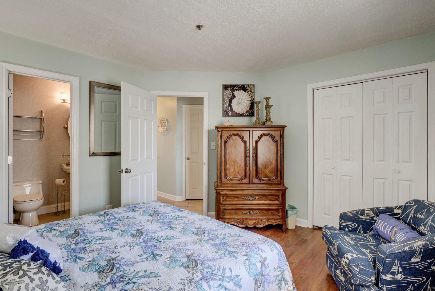 Une chambre à coucher avec un lit, une commode, une chaise et une salle de bain visible à travers une porte ouverte.