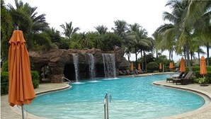 A swimming pool with a waterfall feature, surrounded by palm trees and lounge chairs.