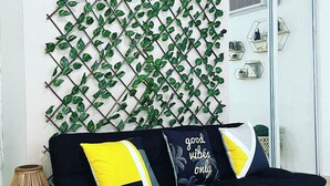 A black sofa with yellow and gray cushions, a woven floor lamp, and a wall-mounted plant grid. - studio apartment near to airport (Carolina)