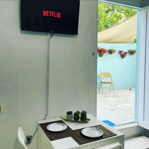 Une pièce avec une table dressée pour deux personnes, une chaise, un téléviseur affichant le logo « NETFLIX », et une vue sur un jardin à l’extérieur.