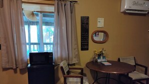Una habitación con una mesa de comedor, cuatro sillas, un refrigerador, una ventana con cortinas, un aire acondicionado de pared y una cita inspiradora enmarcada.