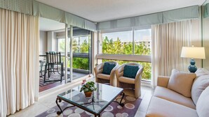 A modern hotel room with a glass coffee table, beige sofa, and two armchairs with teal cushions. A dining area with a table and chairs is visible through the glass door.