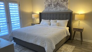 Une chambre avec un grand lit, deux lampes de chevet, une œuvre d’art florale au-dessus du lit et une vue à travers les stores de la fenêtre.