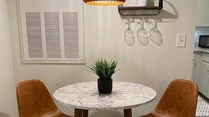 Un coin repas avec une table ronde, deux chaises marron, une plante en pot, un luminaire suspendu et un porte-verres à vin fixé au mur.