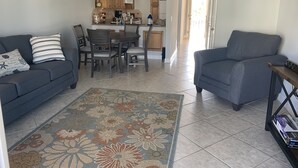 Ein Wohnzimmer mit einer Couch, einem Sessel und einem gemusterten Teppich. Im Hintergrund ist eine Küche mit Holzschränken und einem Essbereich zu sehen.