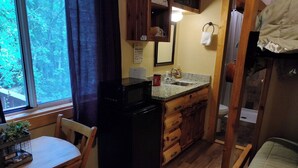 A compact kitchen with a microwave, wooden cabinets, and a window overlooking a forest. - NEW!  Village of Hope - Cabin #12 (Ava)