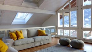 Ein geräumiges Schlafzimmer im Loft-Stil mit geneigter Decke, großen Fenstern, einem grauen Sofa und zwei Poufs.