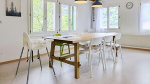 Ein heller, moderner Essbereich mit einem Holztisch, weißen Stühlen, großen Fenstern und einer Uhr an der Wand.