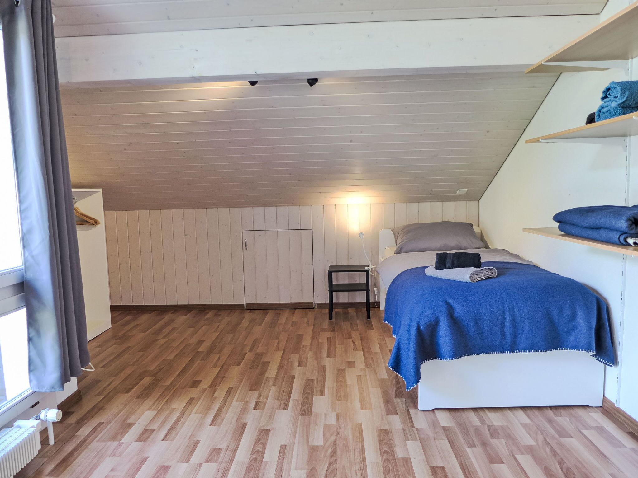 A bedroom with a sloped ceiling, wooden flooring, a bed with a blue blanket, a bedside table, and a shelf with folded towels.