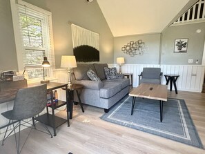 A living room with a grey sofa, a coffee table, a dining table, and a chair. There is a staircase in the background.