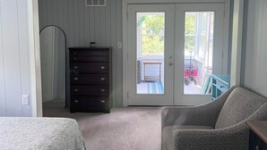 A bedroom with a bed, a dresser, a patterned armchair, and a sliding glass door leading to a balcony.