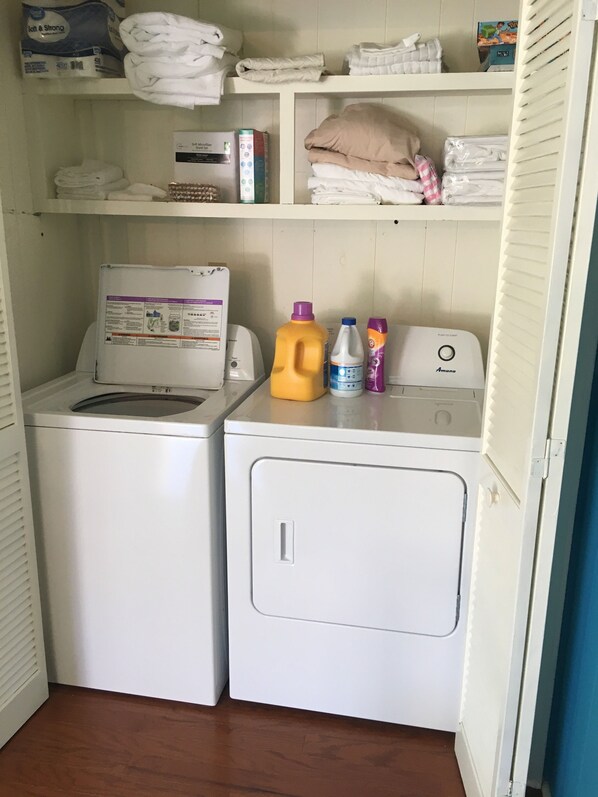 Zona de lavandería con lavadora y secadora, estantes con toallas dobladas y una caja de detergente.