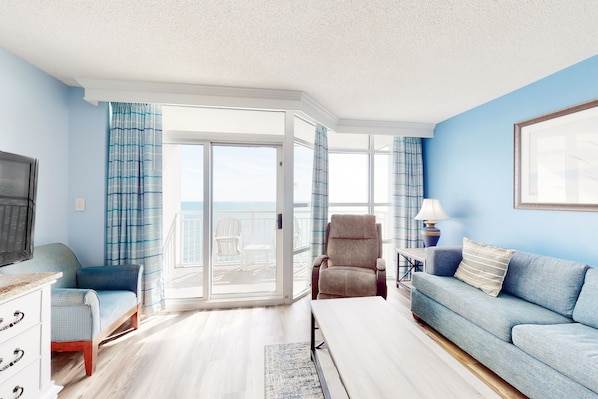 A living room with a blue sofa, a beige armchair, a TV, and a view of the ocean through a sliding glass door.