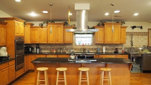 A kitchen with wooden cabinets, a stainless steel range hood, a large island with stools, and a tiled backsplash. - NEW!  Village of Hope - All 6 Cabins (Ava)