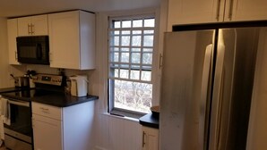 Eine kompakte Küche mit weißen Schränken, einem Edelstahl-Kühlschrank, einer Mikrowelle, einem Toaster und einem Fenster mit Blick auf Bäume.