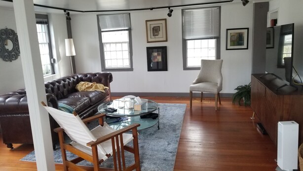 Ein Wohnzimmer mit braunem Ledersofa, einem weißen Sessel, einem Glastisch und einem Holznachttisch. Zwei Fenster mit Jalousien, ein gerahmtes Bild an der Wand und ein Fernseher auf einer Holzkommode.