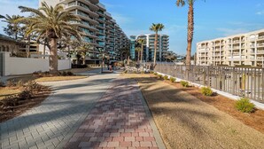 A modern hotel with multiple floors, a paved walkway, palm trees, and a fenced area.
