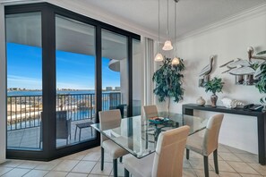A modern dining area with a glass table and chairs, a balcony with a view of the water, and decorative wall art. - Lighthouse on the Bay 2302 | Lazy River | Boat Slip | My Beach Getaways (Orange Beach)