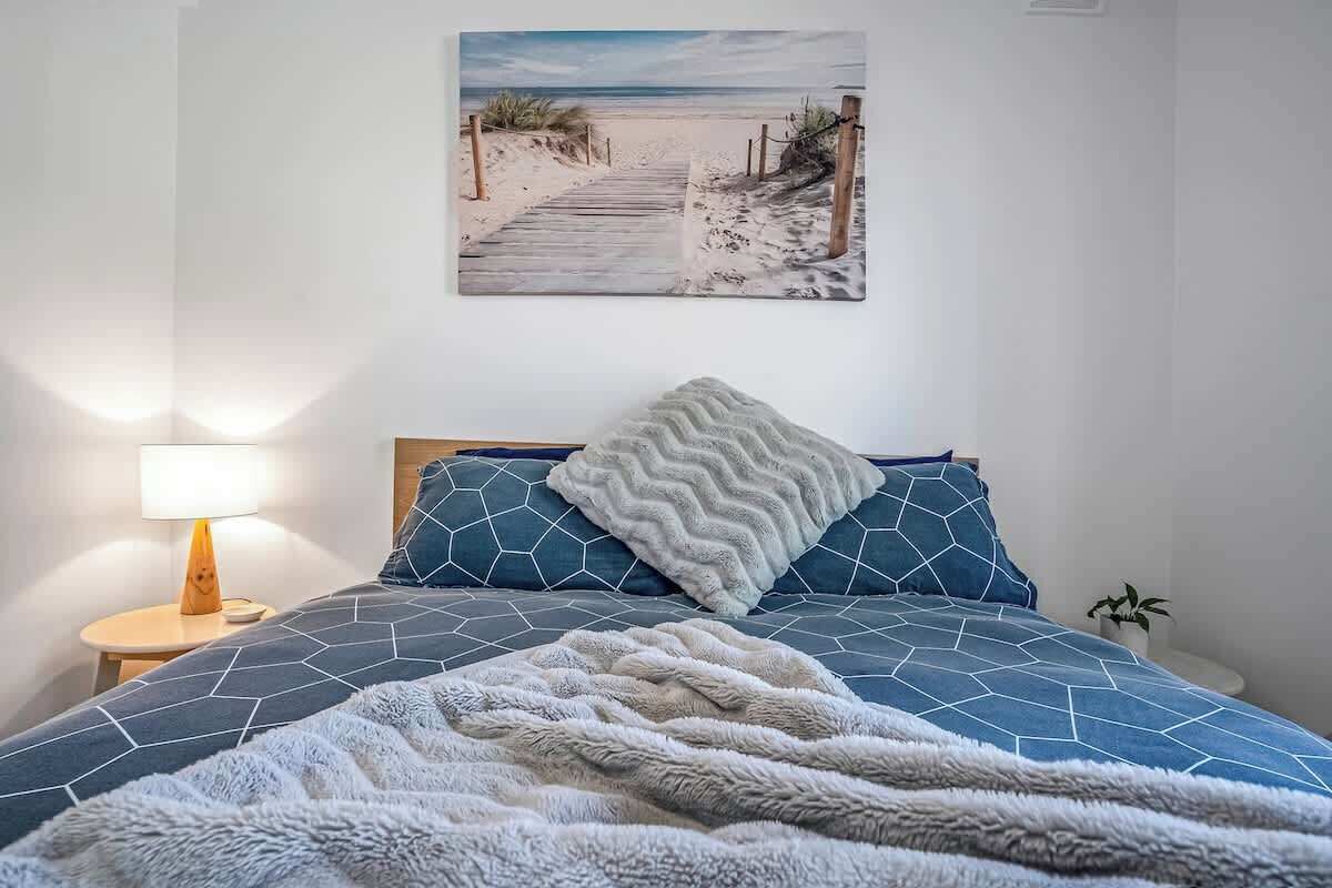 Une chambre à coucher comprenant un lit, une table de chevet, une lampe, un tableau représentant un sentier de plage accroché au mur, et une plante en pot.