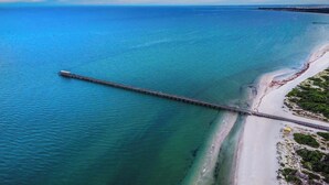 An aerial view of a coastal area with a long pier extending into the water, a sandy beach, and a clear sky. - Beachfront self contained upstairs apartment (Semaphore)