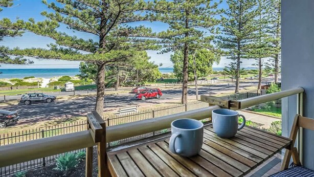 A balcony with a wooden table and two mugs, overlooking a beach with pine trees and a parking area with cars. - Beachfront self contained upstairs apartment (Semaphore)