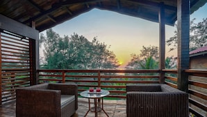 A covered outdoor seating area with wicker furniture and a table with drinks, overlooking a sunset and trees. - Aqua Marina Beach Cottages (Candolim)