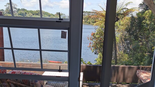 A room with a view of a lake, a wicker chair, and a door leading outside. - With the lake on your doorstep, BEACH 2 minutes away and river swimming hole . (Opunake)