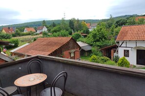 A rooftop terrace with a table and chairs, overlooking a village with traditional houses and a hill in the background.