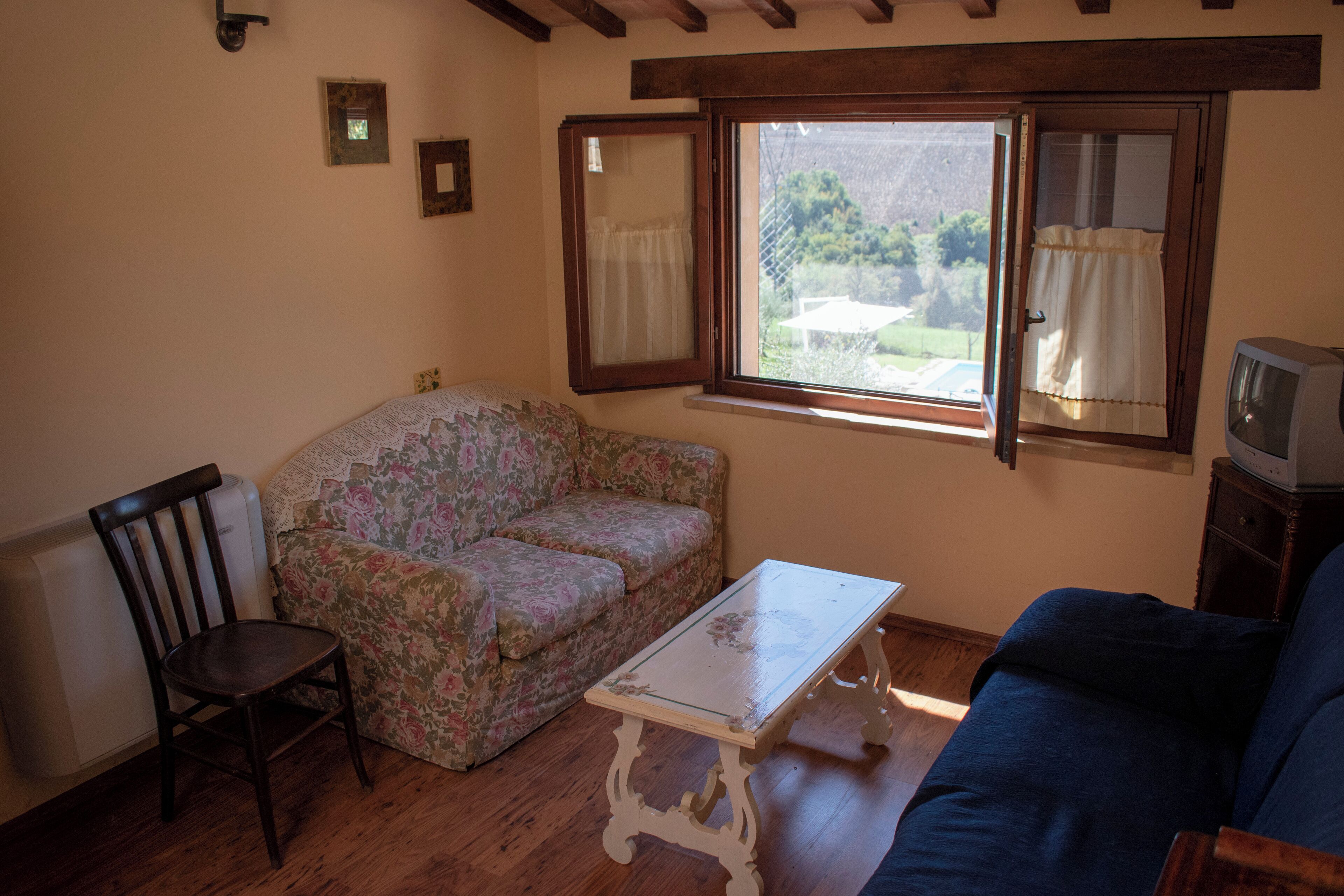 A cozy living room with a floral sofa, a wooden chair, a small white table, and a television on a wooden stand.
