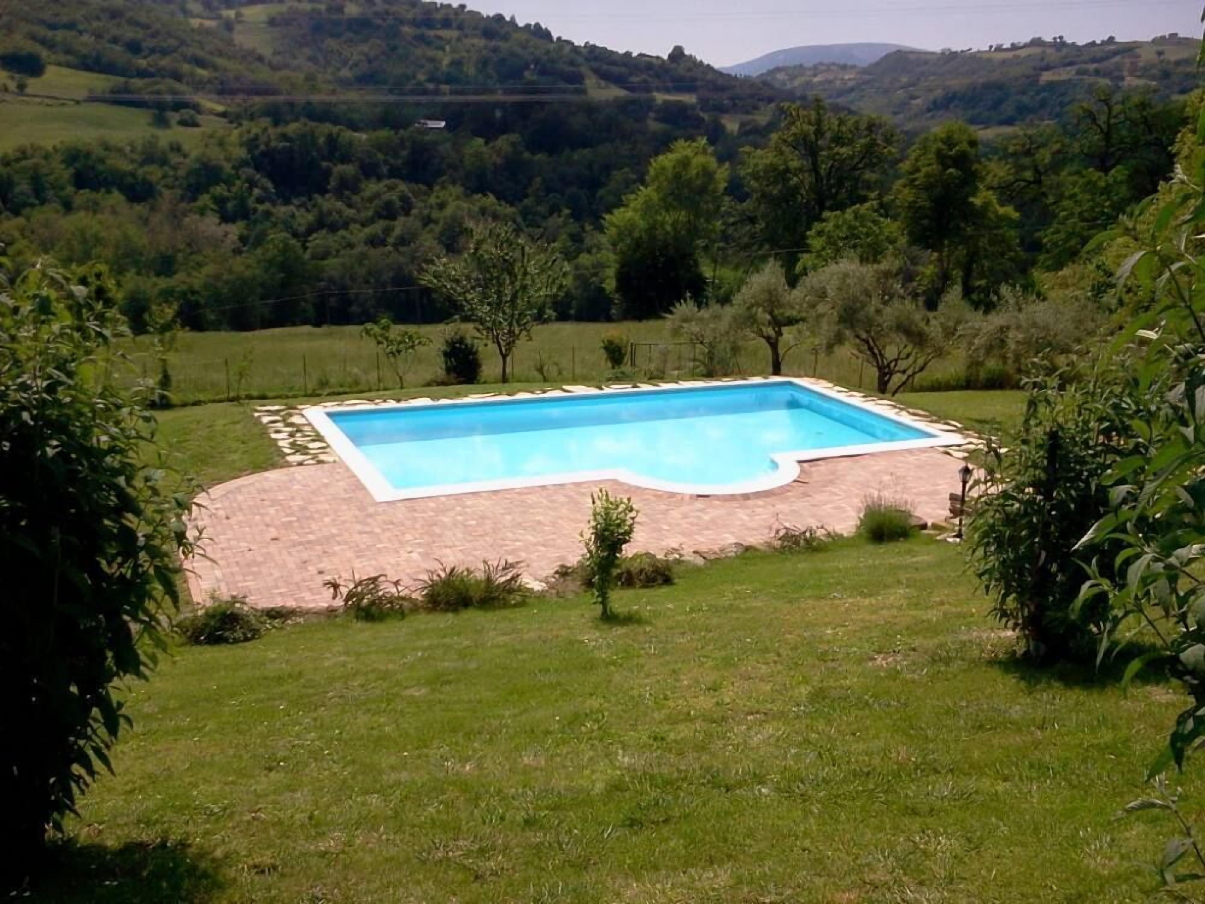 Une piscine rectangulaire entourée d’une terrasse dallée, elle-même bordée de pelouse et d’arbres, le tout situé dans un paysage vallonné.