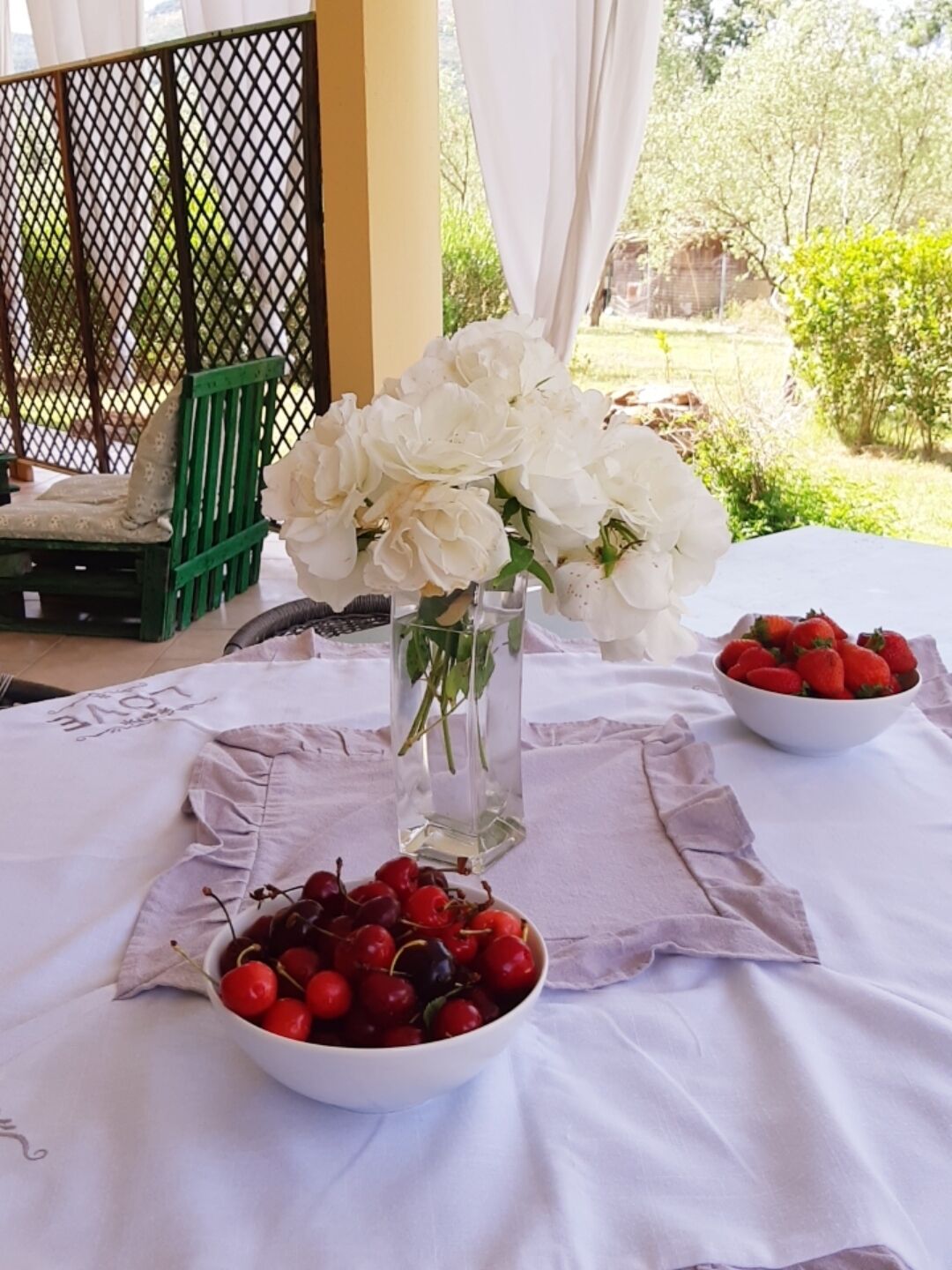 A table with a white tablecloth, a vase of white flowers, a bowl of cherries, and a bowl of strawberries.