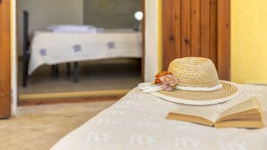 Une pièce avec un lit recouvert d’un drap blanc, une porte en bois et une table sur laquelle se trouvent un chapeau de paille et un livre ouvert.