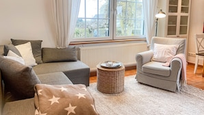A cozy living room with a grey sofa, a checkered armchair, a round wicker ottoman, and a view through a large window. - Ferienhaus Wolff, Deutschland (Barth)