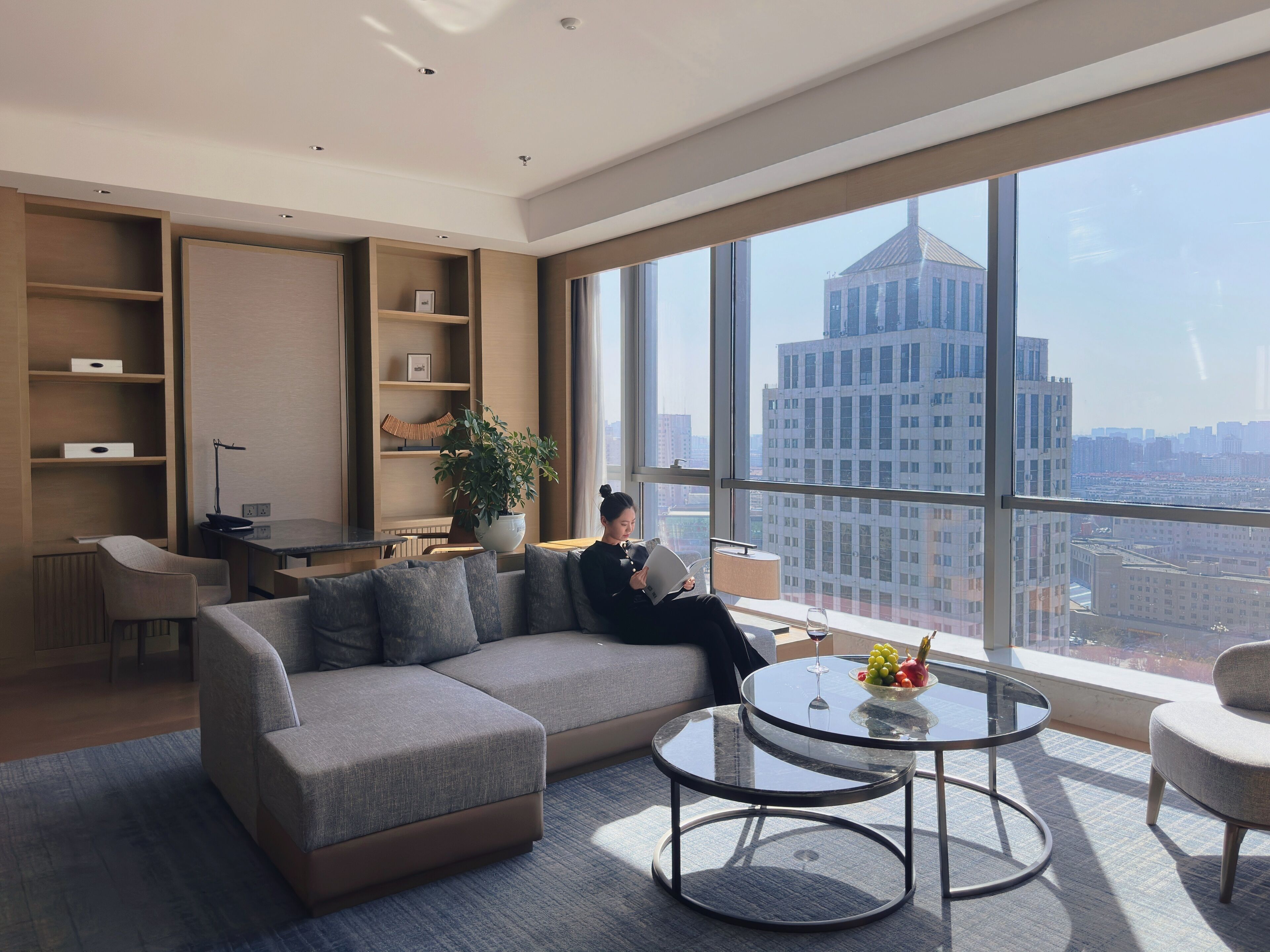 A modern living room with a sofa, coffee table, and a large window offering a city view.