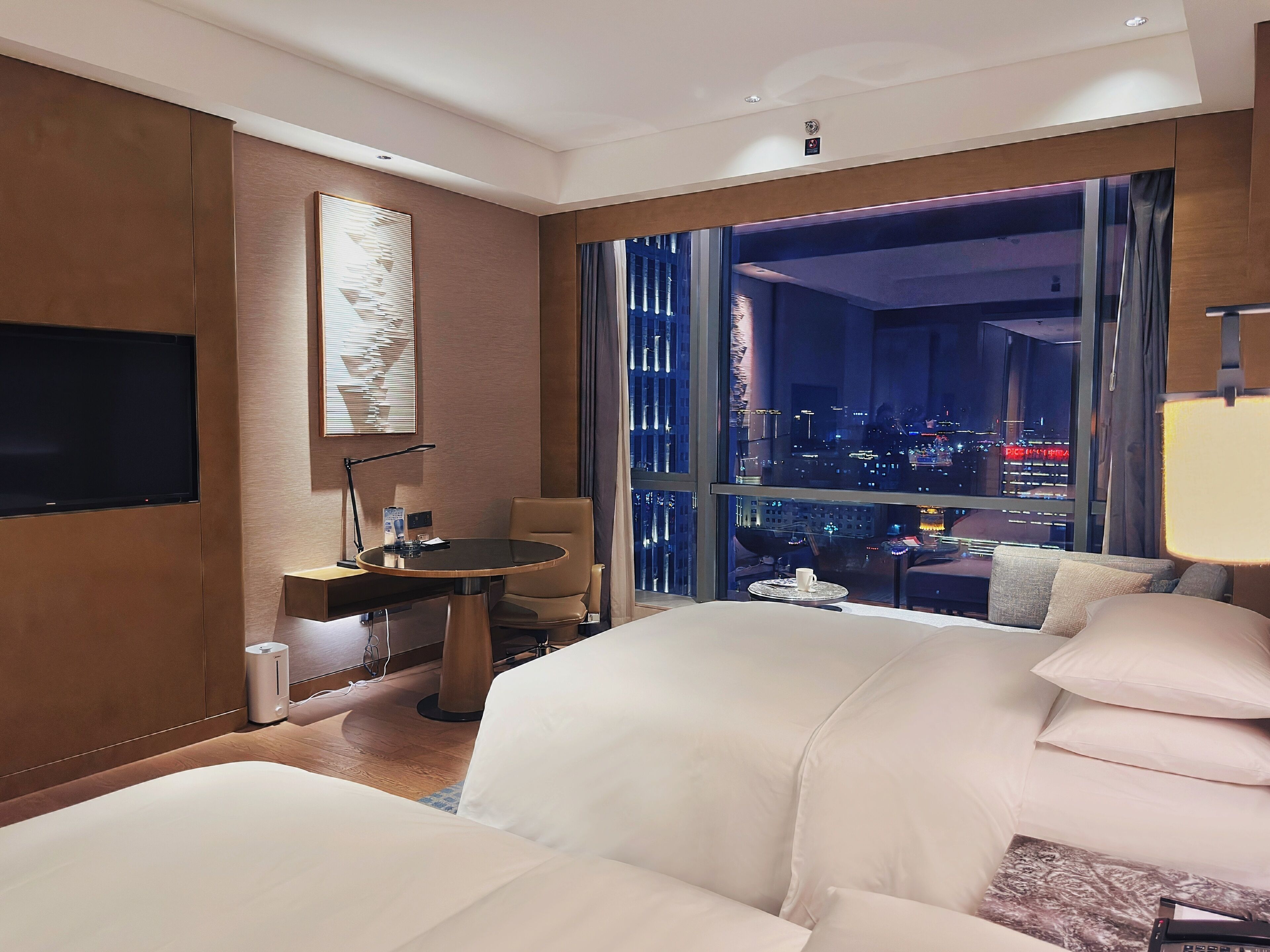 Quarto de hotel com cama, escrivaninha, cadeira, mesa e uma janela grande com vista para a cidade.