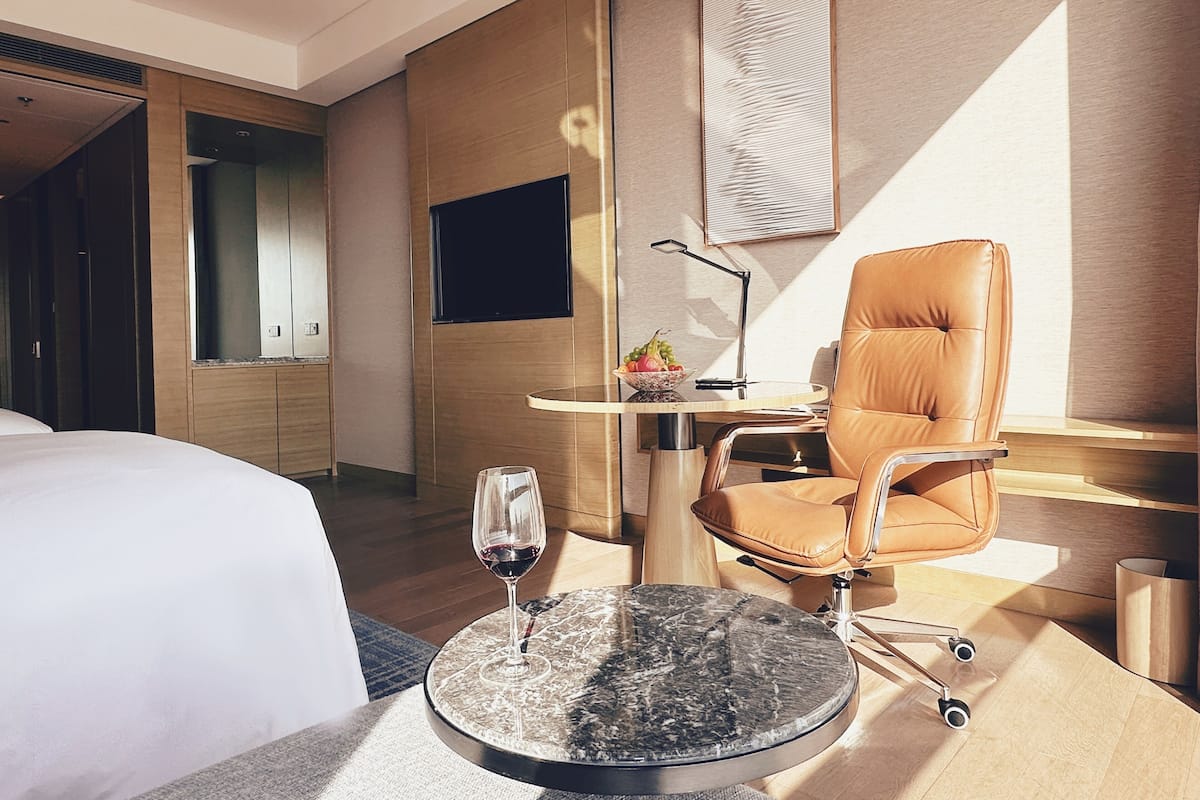 Habitación de hotel con cama, una mesa redonda con una copa de vino, un sillón de cuero, un televisor y una obra de arte colgada en la pared.