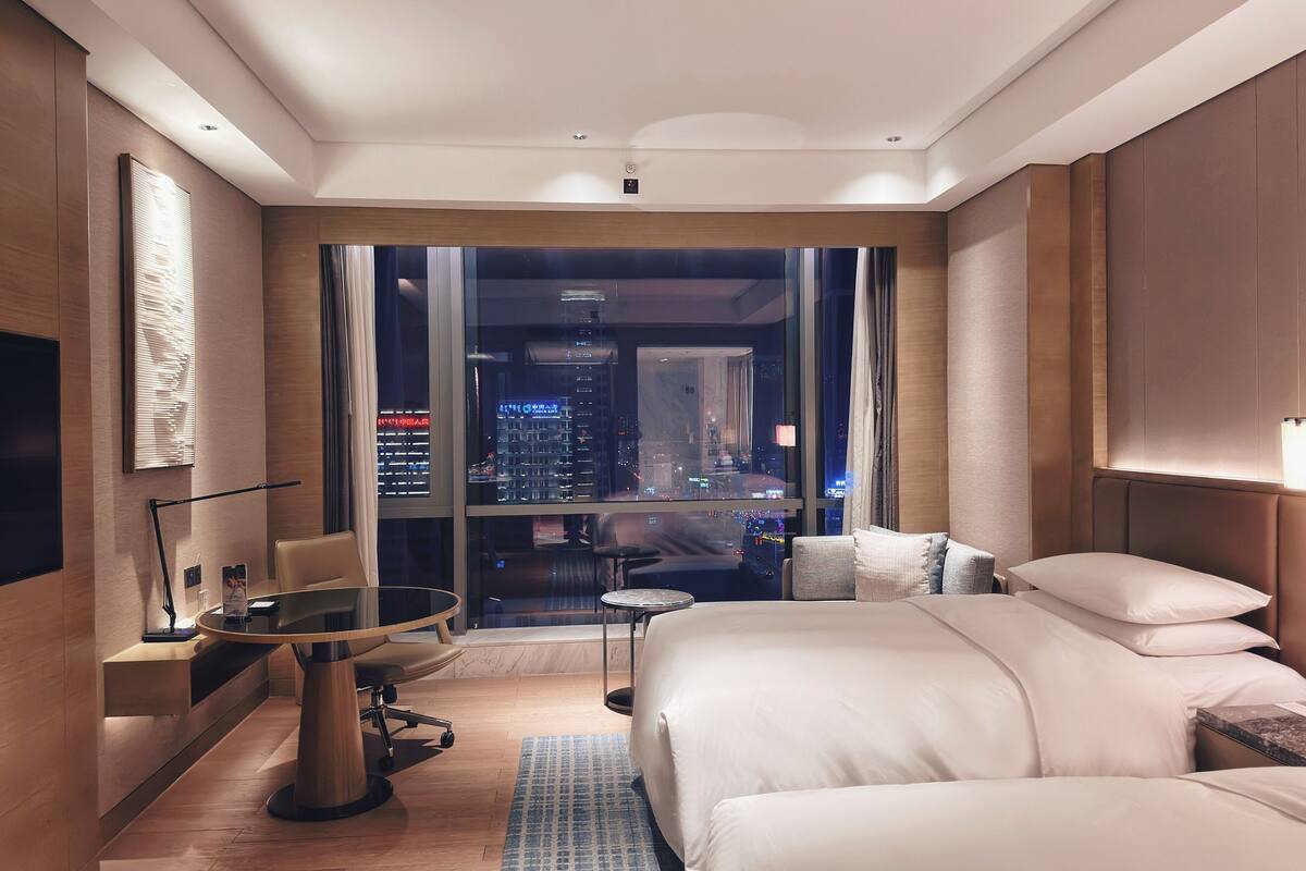 Habitación de hotel moderna con una cama grande, un escritorio, una silla y vistas de un paisaje urbano a través de amplias ventanas.