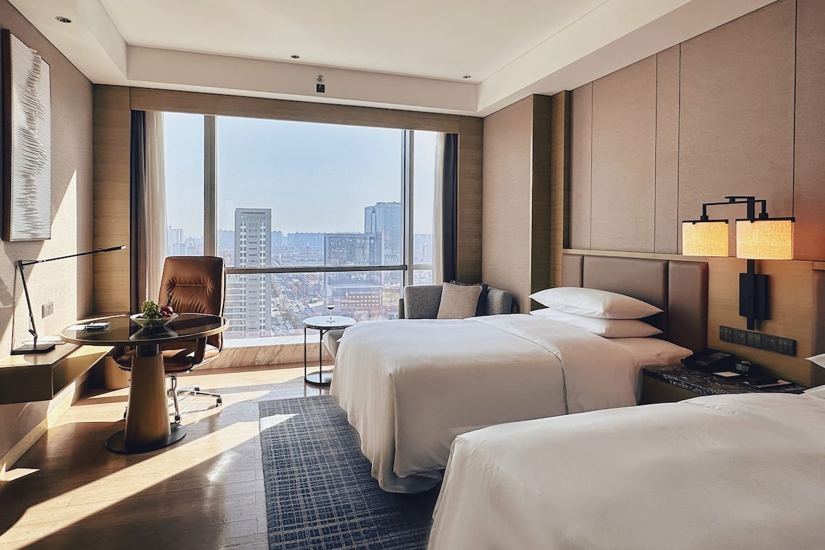 Habitación de hotel con un ventanal grande, dos camas, un escritorio con silla, una lámpara y vistas a la ciudad.