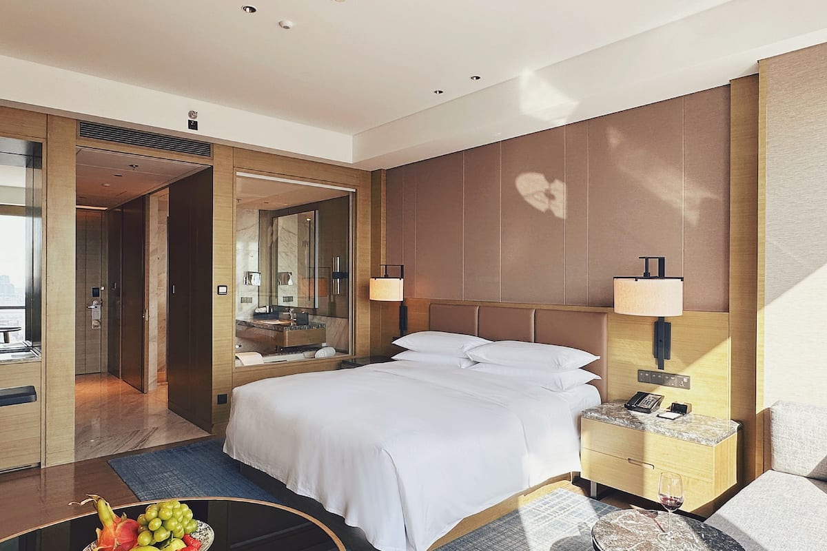 Habitación de hotel con cama, mesita de noche, lámpara y un frutero sobre una mesa.