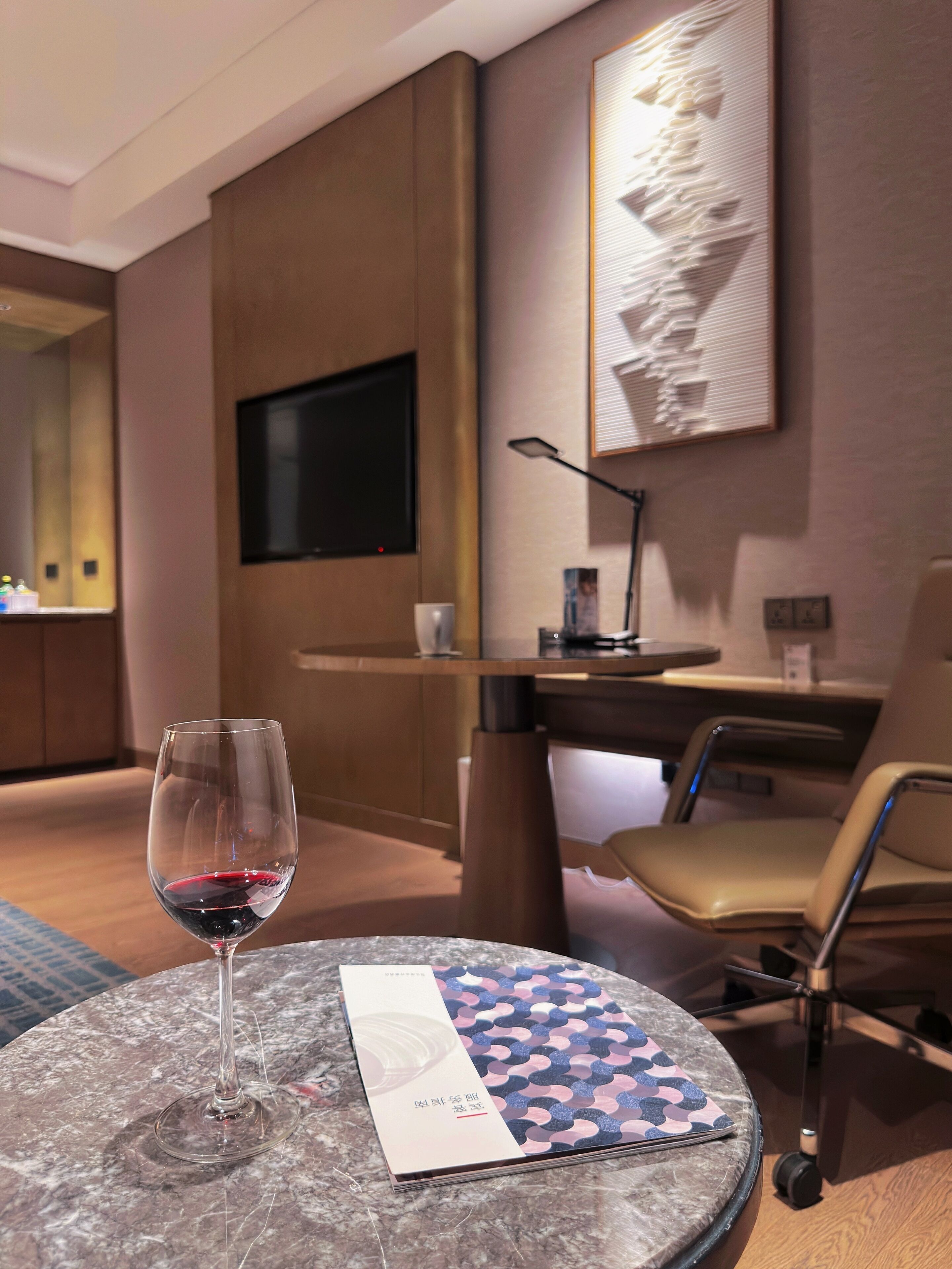 A hotel room with a round table, a glass of red wine, a chair, a desk, a TV, and a wall-mounted artwork.