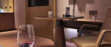 A hotel room with a round table, a glass of red wine, a chair, a desk, a TV, and a wall-mounted artwork.