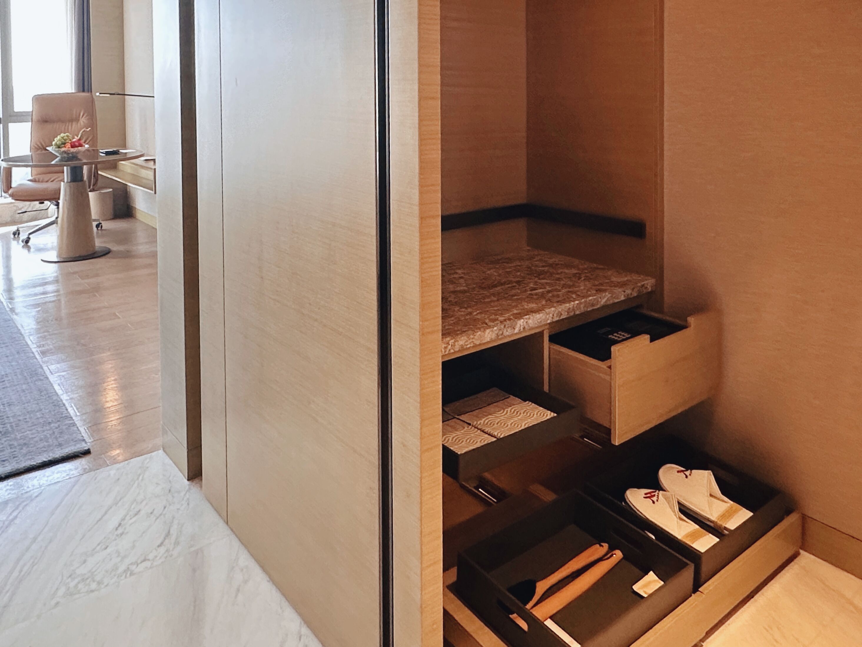 Una habitación de hotel moderna con suelo de mármol, un separador de vidrio, un tocador de madera con cajones y una zona de estar con una silla y una mesa.