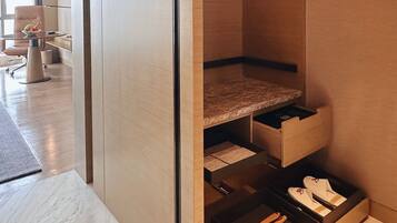 Una habitación de hotel moderna con suelo de mármol, un separador de vidrio, un tocador de madera con cajones y una zona de estar con una silla y una mesa.