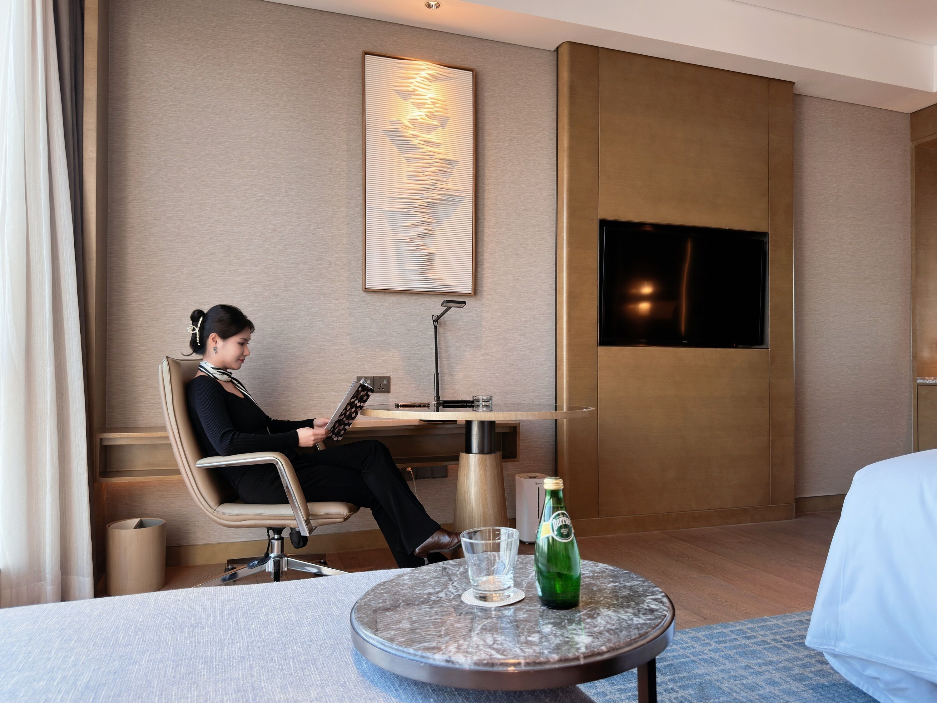 A hotel room with a bed, a round table with a bottle and glass, a desk with a chair, a lamp, and a wall-mounted TV.