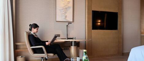 A hotel room with a bed, a round table with a bottle and glass, a desk with a chair, a lamp, and a wall-mounted TV.