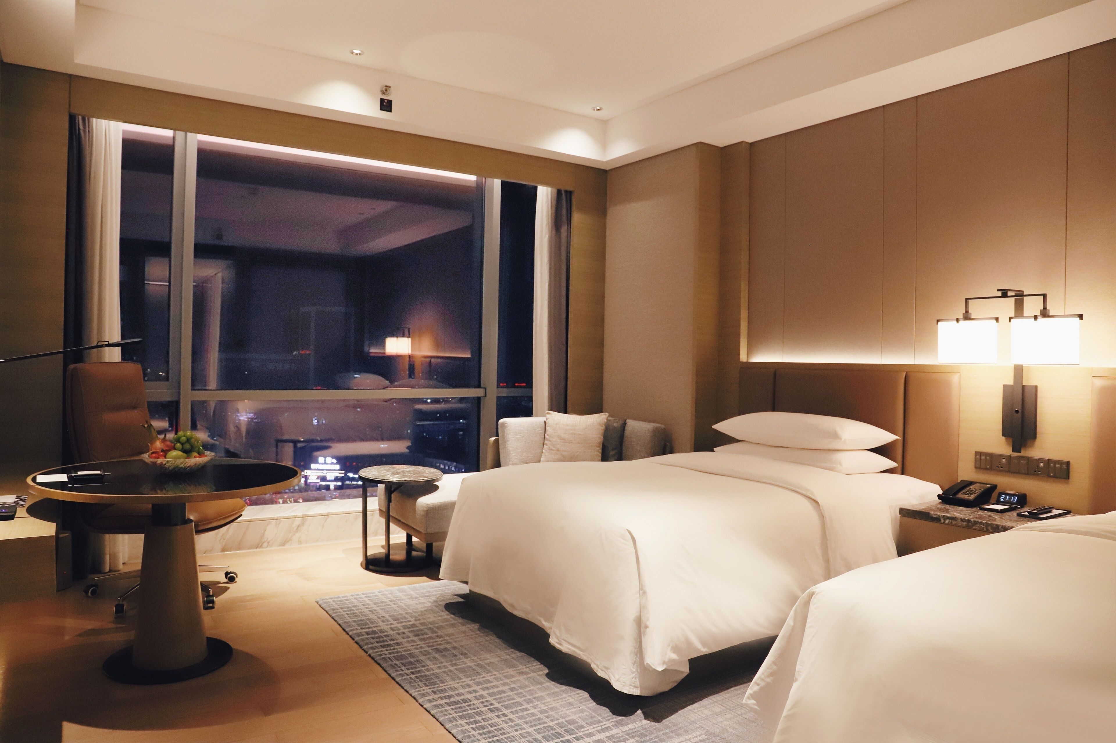 A hotel room with two beds, a dining table with fruit, a small side table, and a large window with a view.