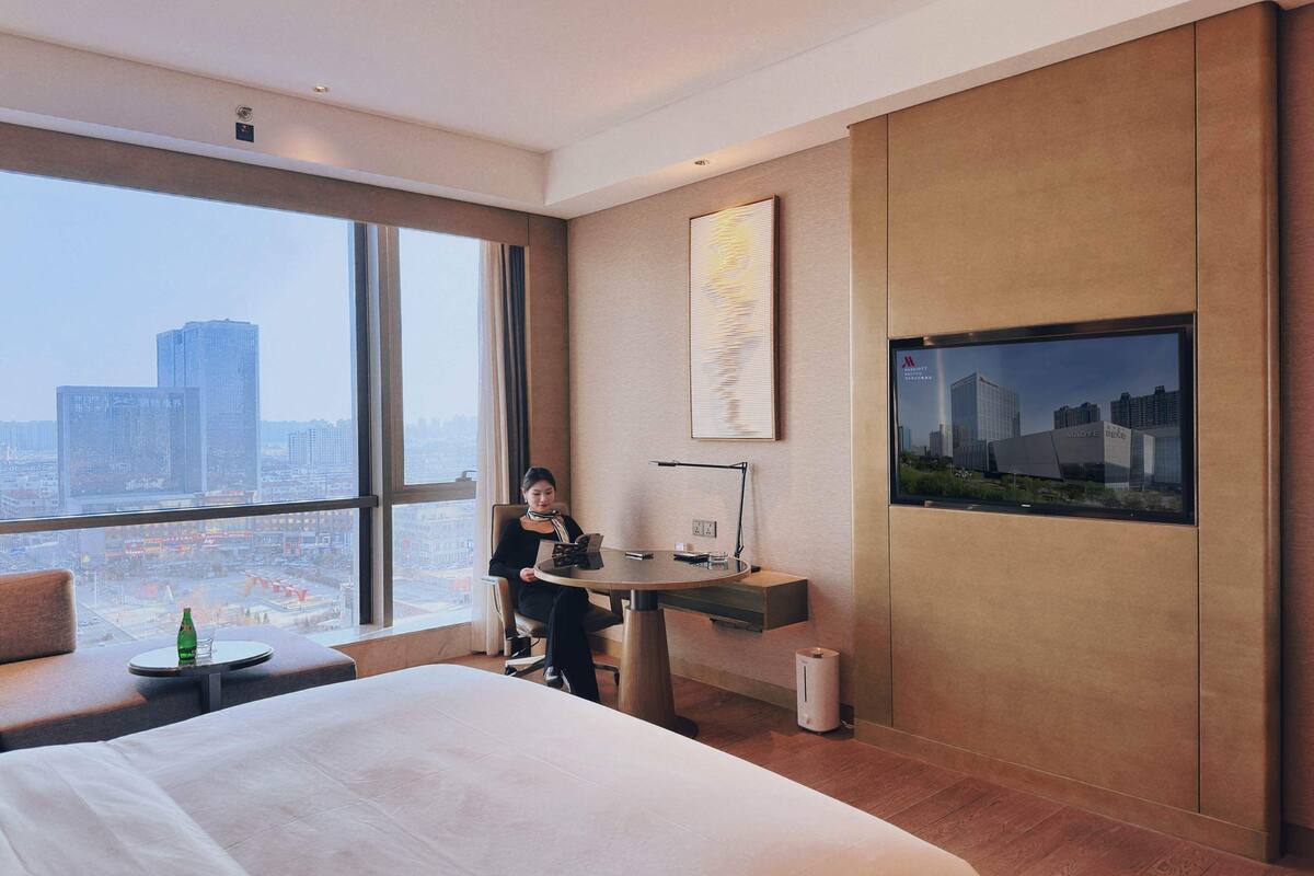 Habitación de hotel con una cama grande, un escritorio, una silla, una mesita y un ventanal con vistas a la ciudad.
